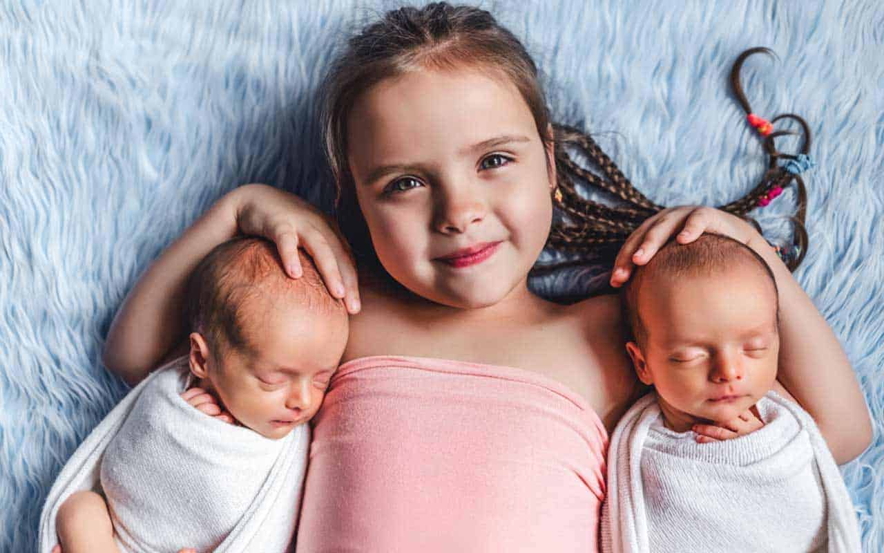 A happy sister lays between her twin newborn siblings