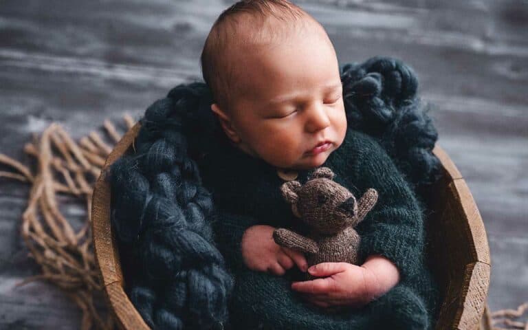 A newborn baby sleeps contently in a basket holding a teddy bear | Career advice for infant care